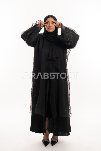 المعاناة من ضعف البصر، التركيز بالنظر على شيء معين، محاولة الرؤية بوضوح، بورتريه لامرأة عربية خليجية اماراتية محجبة ترتدي عباءة سوداء يرفع يديها على شكل نظارات بجانب عينيها، شابة سعودية انيقة، صورة لكامل الجسم، خلفية بيضاء