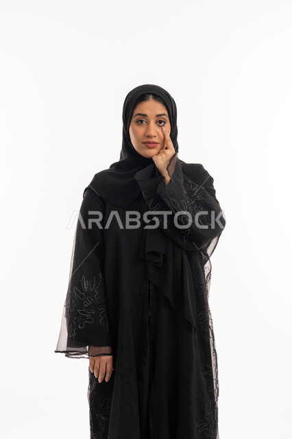 التركيز بالنظر على شيء معين، محاولة الرؤية بوضوح، المعاناة من ضعف البصر، بورتريه لامرأة عربية خليجية اماراتية محجبة ترتدي عباءة سوداء ترفع يدها إلى عينها، شابة سعودية انيقة، خلفية بيضاء