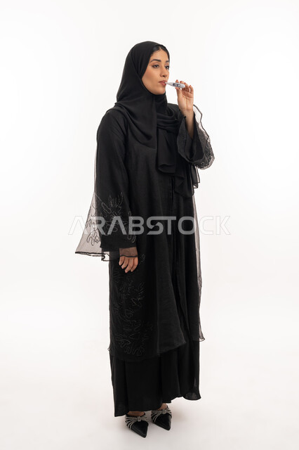 وضع خطة علاجية، الحاجة للمساعدة وزيارة الطبيب، الإصابة بالحمى والتعب، الإحساس بالمرض والإرهاق، بورتريه من الجانب لامرأة عربية خليجية اماراتية محجبة ترتدي عباءة سوداء تقوم بقياس حرارتها باستخدام الميزان الرقمي، شابة سعودية مريضة، صورة لكامل الجسم، خلفية بيضاء