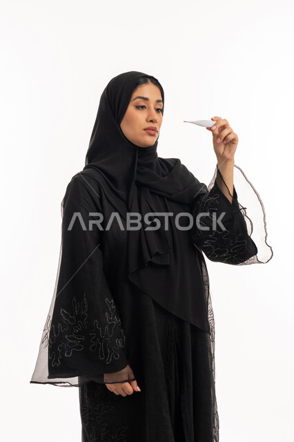 اتباع إجراءات السلامة والوقاية، الإحساس بالمرض والإرهاق، وضع خطة علاجية، الحاجة للمساعدة وزيارة الطبيب، الإصابة بالحمى والتعب، بورتريه من الجانب لامرأة عربية خليجية اماراتية محجبة ترتدي عباءة سوداء تحمل بيدها ميزان حرارة رقمي، شابة سعودية مريضة، خلفية بيضاء