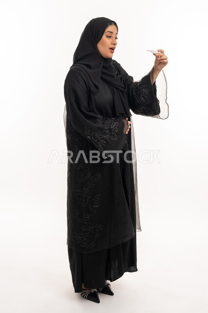 وضع خطة علاجية، الحاجة للمساعدة وزيارة الطبيب، الإصابة بالحمى والتعب، الإحساس بالمرض والإرهاق، بورتريه من الجانب لامرأة عربية خليجية اماراتية محجبة ترتدي عباءة سوداء تحمل بيدها ميزان حرارة رقمي، شابة سعودية مريضة، صورة لكامل الجسم، خلفية بيضاء