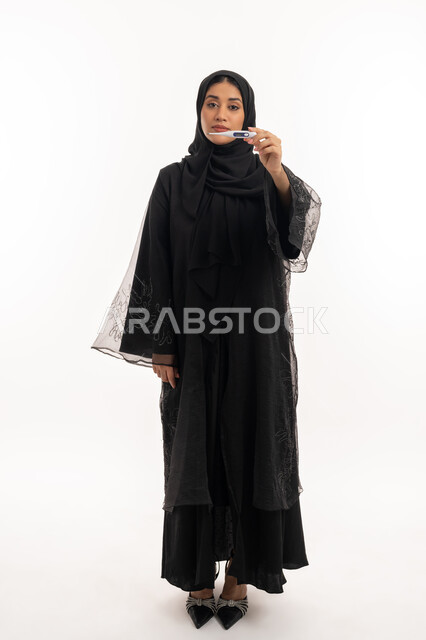 الإصابة بالحمى والتعب، وضع خطة علاجية، الحاجة للمساعدة وزيارة الطبيب، الإحساس بالمرض والإرهاق، بورتريه  لامرأة عربية خليجية اماراتية محجبة ترتدي عباءة سوداء تحمل بيدها الميزان الرقمي، شابة سعودية مريضة، صورة لكامل الجسم، خلفية بيضاء