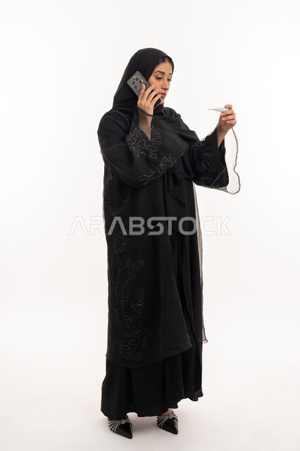 إجراء مكالمة صوتية عبر الهاتف المحمول, الإحساس بالمرض والإرهاق، الإصابة بالحمى والتعب، بورتريه من الجانب لامرأة عربية خليجية اماراتية محجبة تقوم بقياس حرارتها باستخدام الميزان الرقمي، الحاجة لزيارة الطبيب، شابة سعودية مريضة، صورة لكامل الجسم، خلفية بيضاء