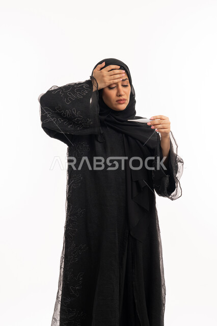 الحاجة للمساعدة وزيارة الطبيب، الإصابة بالحمى والتعب، الإحساس بالمرض والإرهاق، بورتريه  لامرأة عربية خليجية اماراتية محجبة ترتدي عباءة سوداء تضع يدها على جبينها تقوم بقياس حرارتها باستخدام الميزان الرقمي، وضع خطة علاجية، شابة سعودية مريضة، خلفية بيضاء