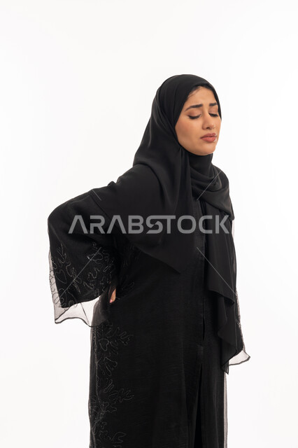 الشعور بالمرض والإرهاق، تعابير الألم ووجع الظهر، بورتريه من الجانب لامرأة عربية خليجية اماراتية محجبة ترتدي عباءة سوداء تضع يدها على ظهرها بايماءات التعب، الحاجة للعلاج والاستشارة الطبية، شابة سعودية مريضة، خلفية بيضاء