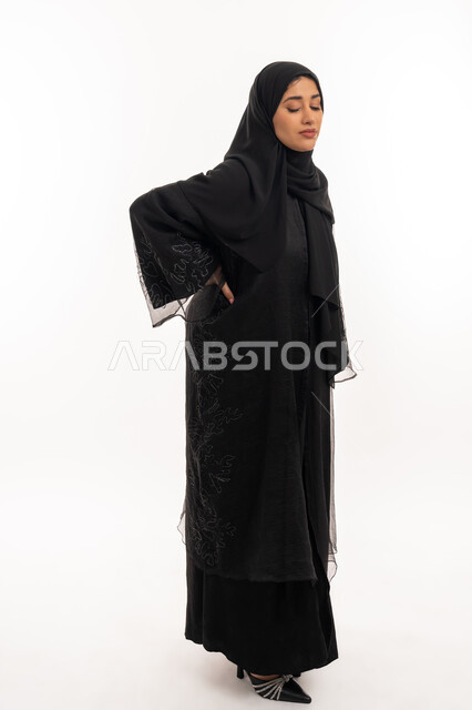 تعابير الألم ووجع الظهر، الشعور بالمرض والإرهاق، بورتريه من الجانب لامرأة عربية خليجية اماراتية محجبة ترتدي عباءة سوداء تضع يدها على ظهرها بايماءات التعب، الحاجة للعلاج والاستشارة الطبية، شابة سعودية مريضة، صورة لكامل الجسم، خلفية بيضاء