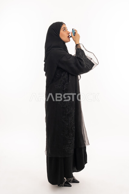شابة سعودية مريضة، المحافظة على ممر هوائي منتظم، مشاكل في الجهاز التنفسي، بورتريه من الجانب لامرأة عربية خليجية اماراتية محجبة ترتدي عباءة سوداء تحمل بيدها جهاز الرذاذ الخاص بمرض الربو تقوم باستنشاق أدوية الكحة، صورة الجسم بالطول الكامل، خلفية بيضاء