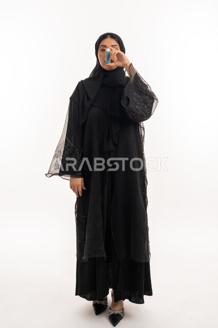 Respiratory problems, portrait of veiled Gulf Arab Emirati woman wearing black abaya holding asthma nebulizer inhaling cough medicine, maintaining regular airway, young Saudi patient, full body image, white background