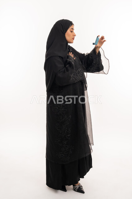 أجهزة استنشاق المخصصة للمرضى، مشاكل في الجهاز التنفسي، بورتريه من الجانب لامرأة عربية خليجية اماراتية محجبة ترتدي عباءة سوداء تحمل بيدها جهاز الرذاذ الخاص بمرض الربو، المحافظة على ممر هوائي منتظم، شابة سعودية مريضة، صورة الجسم بالطول الكامل، خلفية بيضاء