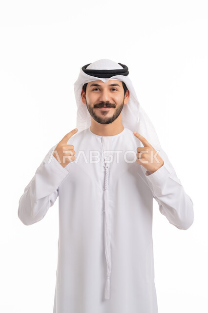الأناقة والرجولة والاهتمام بالمظهر الخارجي, الوقوف والنظر إلى الكاميرا بإيماءات السرور, بورتريه لرجل عربي خليجي إماراتي يرتدي الكندورة والغترة الاماراتية يشير إلى وجهه بسعادة, مفهوم الثقة بالنفس, خلفية بيضاء