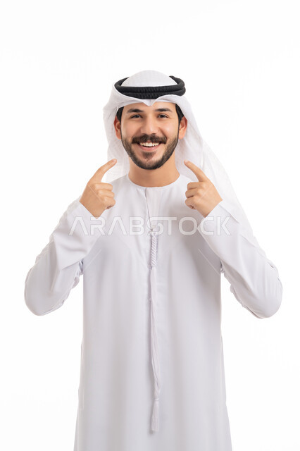 مفهوم الثقة بالنفس, النظر إلى الكاميرا بإيماءات السرور, استخدام قشور فينير من الخزف, ابتسامة بيضاء ناصعه، أهمية متابعة طبيب الاسنان، بورتريه لشاب عربي خليجي إماراتي مبتسم يرتدي الكندورة والغترة يشير بيديه إلى أسنانه بسعادة, خلفية بيضاء