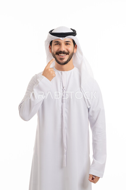 النظر إلى الكاميرا بإيماءات السرور, استخدام قشور فينير من الخزف, ابتسامة بيضاء ناصعه، أهمية متابعة طبيب الاسنان، العناية والتجميل والتبيض، بورتريه لشاب عربي خليجي إماراتي مبتسم يرتدي الكندورة والغترة يشير بيديه إلى أسنانه بسعادة, خلفية بيضاء