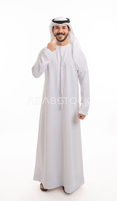 الوقوف باستقامة والنظر إلى الكاميرا, استخدام قشور فينير من الخزف لصنع ابتسامة بيضاء ناصعه، أهمية متابعة طبيب الاسنان، العناية والتجميل والتبيض، بورتريه لشاب عربي خليجي إماراتي مبتسم يرتدي الكندورة والغترة يشير بيديه إلى أسنانه, صورة الجسم بالكامل, خلفية بيضاء