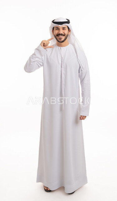 استخدام قشور فينير من الخزف لصنع ابتسامة بيضاء ناصعه، أهمية متابعة طبيب الاسنان، العناية والتجميل والتبيض، بورتريه لشاب عربي خليجي إماراتي مبتسم يرتدي الكندورة والغترة ينظر إلى الكاميرا يشير بيديه إلى أسنانه, صورة الجسم بالطول الكامل, خلفية بيضاء