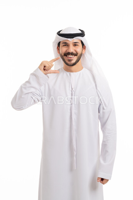 ابتسامة بيضاء ناصعه، أهمية متابعة طبيب الاسنان، العناية والتجميل والتبيض، تعابير السرور باستخدام قشور فينير من الخزف، بورتريه لشاب عربي خليجي إماراتي مبتسم يرتدي الكندورة والغترة ينظر إلى الكاميرا يشير بيديه إلى أسنانه, خلفية بيضاء