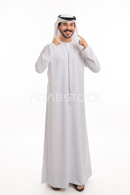 العناية والتجميل والتبيض، ابتسامة بيضاء ناصعه، تعابير السرور باستخدام قشور فينير من الخزف، بورتريه لشاب عربي خليجي إماراتي مبتسم يرتدي الكندورة والغترة ينظر إلى الكاميرا يشير بيديه إلى أسنانه, أهمية متابعة طبيب الاسنان، صورة الجسم بالطول الكامل, خلفية بيضاء
