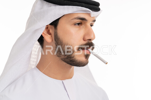 تعاطي مواد ضارة وسامة، عادات سلوكية خاطئة, الإصابه بأمراض الجهاز التنفسي، بورتريه مقرب من الجانب لشاب عربي خليجي اماراتي يرتدي الكندورة والغترة يقوم بسحب نفس عميق من السيجارة، الادمان على التبغ والسجائر، خلفية بيضاء