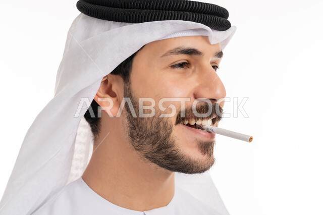 عادات سلوكية خاطئة, الإصابه بأمراض الجهاز التنفسي، بورتريه مقرب من الجانب لشاب عربي خليجي اماراتي مبتسم يرتدي الكندورة والغترة يقوم بسحب نفس عميق من السيجارة، الادمان على التبغ والسجائر، تعاطي مواد ضارة وسامة، النظر إلى الكاميرا بسرور، خلفية بيضاء