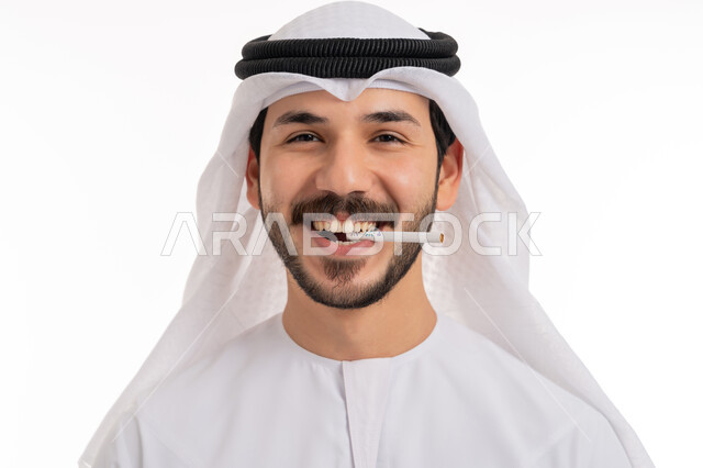 الإصابه بأمراض الجهاز التنفسي، بورتريه مقرب لشاب عربي خليجي اماراتي مبتسم يرتدي الكندورة والغترة يقوم بسحب نفس عميق من السيجارة، الادمان على التبغ والسجائر، تعاطي مواد ضارة وسامة، النظر إلى الكاميرا بسرور, عادات سلوكية خاطئة، خلفية بيضاء