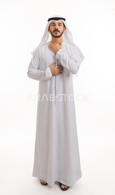 Infection with the Corona virus, looking at the camera with gestures of difficulty breathing, fatigue and pain, having a cold and sore throat, portrait of an Arab Gulf Emirati young man wearing a kandora and ghutra, putting his hands on his throat and feeling suffocated, severe illness and coughing, full-length body photo, white background.