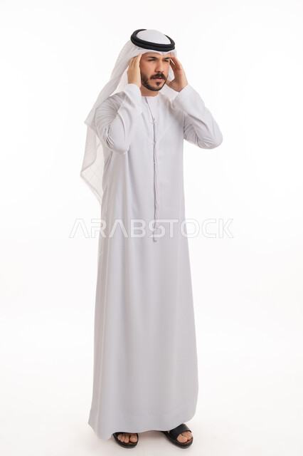 الاحساس بالشقيقية والصداع النصفي, الإرهاق وفقدان التركيز, الشعور بالصداع وألم الرأس, الاجهاد والضغط النفسي, بورتريه لرجل عربي خليجي اماراتي يرتدي الكندورة والغترة يضع يديه على رأسه ويغمض عينيه بإيماءات الألم الشديد, صورة الجسم بالطول الكامل, خلفية بيضاء