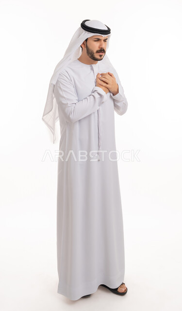 اضطرابات وأمراض قلبية, التعب والإرهاق والضغوطات النفسية, الشعور في زيادة معدل ضربات القلب, الاحساس بذبحه صدريه، بورتريه لشاب عربي خليجي اماراتي يرتدي الكندورة والغترة يضع يده على قلبه ويشعر بحالة سيئة من الوخز والألم, صورة الجسم بالطول الكامل, خلفية بيضاء