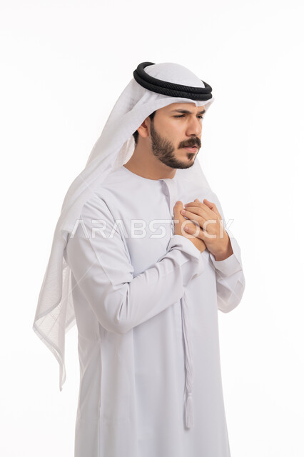 Fatigue, exhaustion, and psychological pressures, heart disorders and diseases, the feeling of an increased heart rate, the feeling of a pounding in his chest, a portrait of an Arab Gulf Emirati young man wearing a kandora and a ghutra, placing his hand on his heart, feeling a bad state of tingling and pain in his heart, white background.