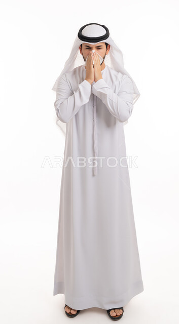 The need for treatment and medicine, feeling sick and cold, allergic rhinitis, using tissues to sneeze, portrait from the side of a young Arab Emirati Gulf man wearing a ghutra and kandora, placing his hands on his face with a sneezing gesture, full-length body portrait, white background