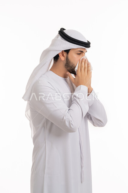 Using paper tissues, feeling sick and having a cold and the need for treatment and medication, allergic rhinitis, portrait from the side of a young Arab Emirati Gulf man wearing a ghutra and kandura, placing his hands on his face with sneezing gestures, white background.