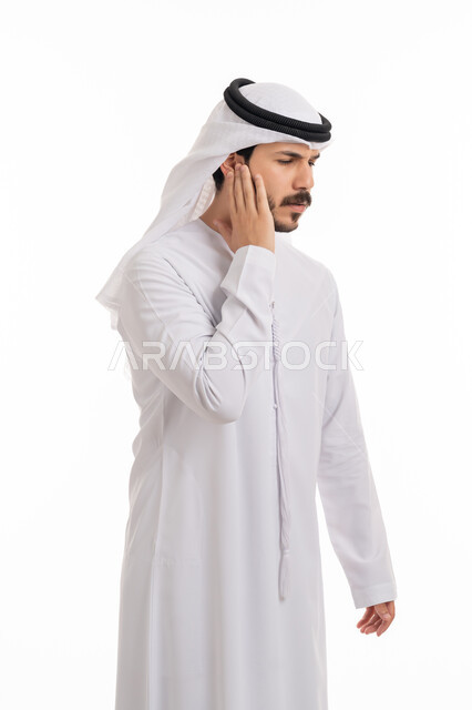 الحاجة للذهاب للطبيب للمساعدة و وضع خطة علاجية مناسبة، حالة اسعافية طارئة، التهاب الأذن الوسطى، بورتريه لشاب عربي خليجي إماراتي يرتدي الكندورة والغترة يضع يده على اذنه بايماءات الألم الشديد, خلفية بيضاء