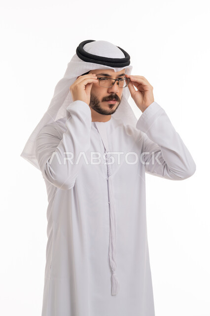 التمعن والتدقيق بشيء ما, مفهوم طول النظر وقصره, بورتريه لشاب عربي خليجي اماراتي يرتدي الكندورة والغترة الاماراتية يمسك النظارة الطبية بإيماءات التركيز, تحسين الرؤية ومحاولة تعديل البصر، خلفية بيضاء
