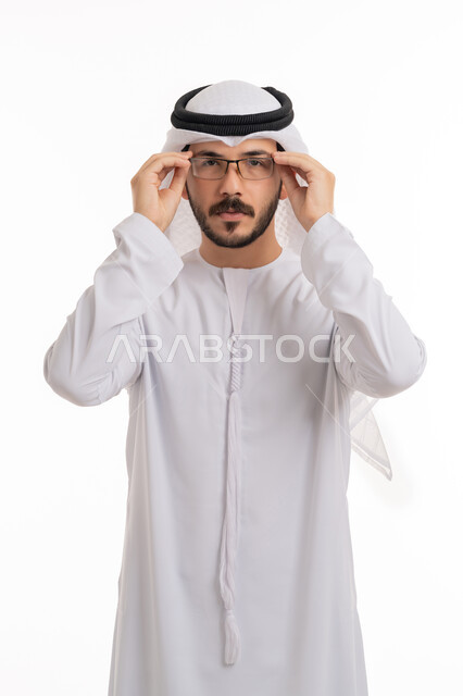 تحسين الرؤية ومحاولة تعديل البصر، التمعن والتدقيق بشيء ما, مفهوم طول النظر وقصره, بورتريه لشاب عربي خليجي اماراتي يرتدي الكندورة والغترة الاماراتية يمسك النظارة الطبية بإيماءات التركيز, خلفية بيضاء