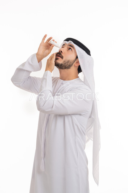 Treating the eye using the necessary drops, the need to examine cases of optic neuritis, a portrait of an Emirati Gulf Arab man wearing a kandura and a ghutra, applying eye drops, high eye pressure, a burning sensation and redness of the eyes, a white background.