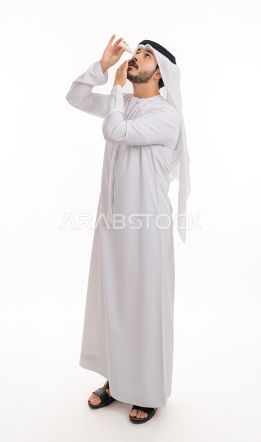 الحاجة للفحص وعلاج حالات التهاب العصب البصري، بورتريه لرجل عربي خليجي اماراتي يرتدي الكندورة والغترة يقوم بوضع قطرة عينية, ارتفاع ضغط العين, الاحساس بالحرقة و احمرار العيون، صورة الجسم بالطول الكامل, خلفية بيضاء