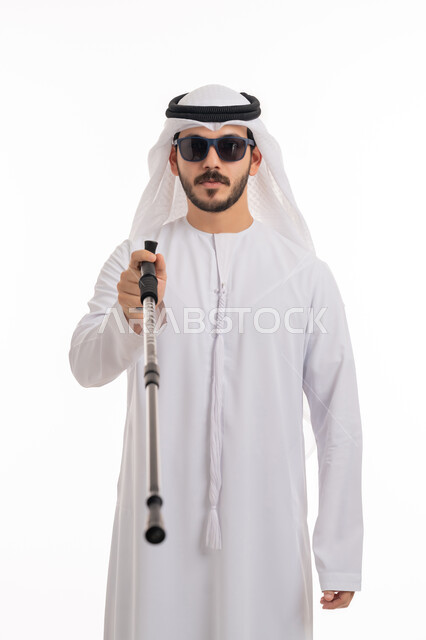 Using a cane to help with balance and movement without stumbling, a portrait of a blind Arab Gulf Emirati man wearing a ghutra and a kandura, wearing sunglasses and pointing at something, providing safe roads for the blind in the Emirates, tools and equipment that help blind people, white background.