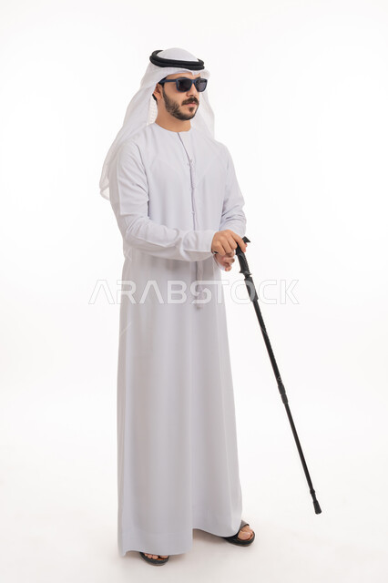 الاعتماد على الدلائل الصوتية المحيطة، التحسس للعقبات وتجنبها أثناء المشي، بورتريه لرجل عربي خليجي إماراتي ضرير يرتدي الكندورة والغترة يضع النظارة الشمسية يمسك العصا لمساعدته في التنقل، صورة الجسم بالطول الكامل, خلفية بيضاء
