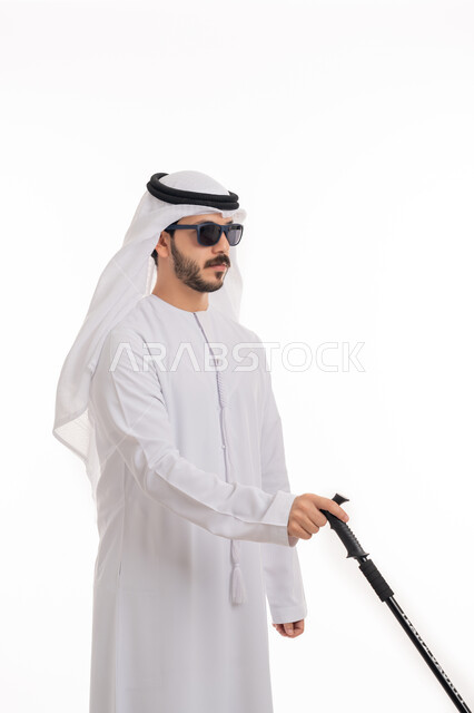 التحسس للعقبات وتجنبها أثناء المشي, الاعتماد على الدلائل الصوتية المحيطة، بورتريه لرجل عربي خليجي إماراتي ضرير يرتدي الكندورة والغترة يضع النظارة الشمسية يمسك العصا لمساعدته في التنقل، خلفية بيضاء