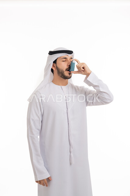 المعاناة من الأزمات التحسسية وضيق التنفس, بورتريه لرجل عربي خليجي اماراتي يرتدي الكندورة والغترة يستخدم  جهاز الرذاذ الخاص بمرض الربو, المحافظة على ممر هوائي منتطم, مشاكل وأمراض صدرية, اجهزة استنشاق خاصة للمرضى, خلفية بيضاء