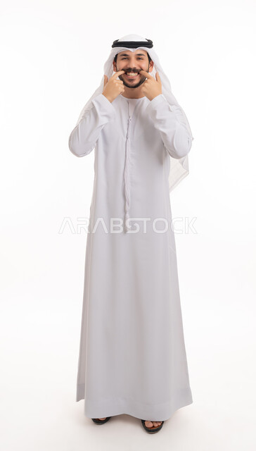 العناية والتجميل والتبيض، ابتسامة بيضاء ناصعه، تعابير السرور باستخدام قشور فينير من الخزف، بورتريه لشاب عربي خليجي إماراتي مبتسم يرتدي الكندورة والغترة ينظر إلى الكاميرا يشير بيديه إلى أسنانه, أهمية متابعة طبيب الاسنان، صورة الجسم بالطول الكامل, خلفية بيضاء