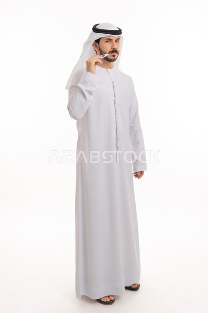 الوقوف بانزعاج والنظر إلى الكاميرا, بورتريه لرجل عربي خليجي اماراتي يرتدي الكندورة والغترة الاماراتية يقوم بقياس درجة حرارته باستخدام الميزان الرقمي, أعراض الاصابة بالحمى والتعب الجسدي, مفهوم الاهتمام بالصحة, صورة الجسم بالطول الكامل, خلفية بيضاء