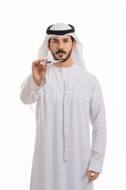 Displaying the digital thermometer, the necessity of having a thermometer at home, a portrait of an Arab Gulf Emirati man wearing a kandora and ghutra, looking at the camera with gestures of fatigue and exhaustion due to illness, a white background.