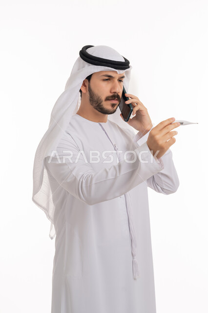 الحاجة إلى المساعدة الطبية, الشعور بالتعب والإرهاق وأعراض الحمى، اجراء مكالمة هاتفية عبر الجوال لاستشارة الطبيب عن العلاج المناسب، بورتريه لشاب عربي خليجي إماراتي يرتدي الكندورة والغترة يمسك بيده جهاز مقياس الحرارة ينظر الى المؤشر, خلفية بيضاء