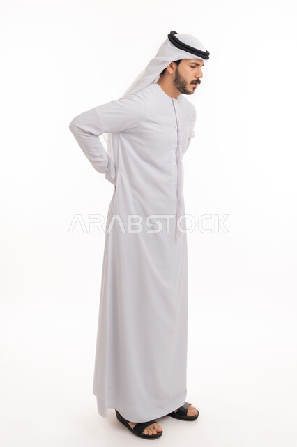 The need for treatment, spine and back pain, feeling of physical aches and pain, fatigue, exhaustion, and feeling sick, a portrait of an Arab Gulf Emirati man wearing a kandura and a ghutra, placing his hand on his back, full-length body portrait, white background.