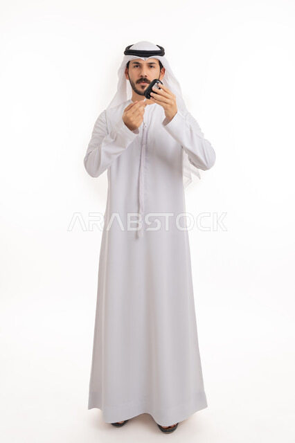 الوقوف باستقامة والنظر إلى الكاميرا بإيماءات التعب, الإصابة بداء السكري، بورتريه لشاب عربي خليجي اماراتي مريض يرتدي الكندورة والغترة الاماراتية يقوم بفحص نسبة السكر في دمه، أجهزة الكترونية حديثة للفحص الطبي السريع، صورة الجسم بالطول الكامل, خلفية بيضاء