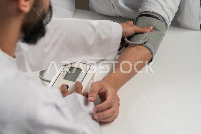 مفهوم الرعاية الصحية والاهتمام بالصحة الجسدية, استخدام الميزان الالكتروني الحديث والمتطور للفحص المنزلي السريع, بورتريه مقرب من الخلف لرجل عربي خليجي اماراتي يرتدي الكندورة الاماراتية يقوم بقياس ضغط الدم للمريض, خلفية رمادية