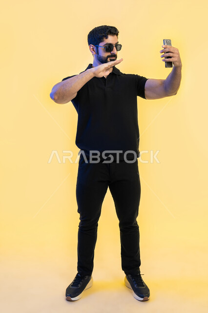 إجراء مكالمة فيديو عبر الجوال، تبادل النقاشات والأحاديث، مفهوم لغة الجسد، بورتريه لشاب عربي خليجي اماراتي يرتدي زي الكاجوال ويضع النضارات الشمسية، التواصل مع الأصدقاء والأقارب على السوشال ميديا بإيماءات السعادة، صورة الجسم بالطول الكامل، خلفية صفراء