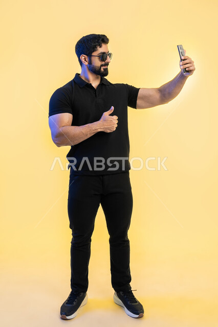 رفع إصبع الابهام نحو الأعلى بعلامة الجودة، إجراء مكالمة فيديو مع الأصدقاء, الاعجاب بآداء التقنية الحديثة، بورتريه لشاب عربي خليجي اماراتي مبتسم يرتدي الزي الكاجوال ويضع النظارات يمسك بيده الهاتف المحمول، صورة الجسم بالطول الكامل، خلفية صفراء