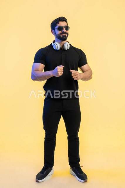 إستخدام سماعة الرأس والتقنيات المتطورة، الاستمتاع في أوقات الفراغ, التفاعل مع الأنغام الوسيقية، مفهوم الإصغاء والإستماع، بورتريه لشاب عربي خليجي اماراتي يرتدي الزي الكاجوال والنظارات الشمسية، صورة الجسم بالطول الكامل، خلفية صفراء