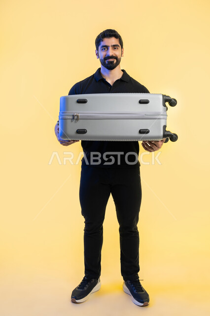 Fun and entertainment during the summer vacation, spending enjoyable times outside the country, portrait of a smiling Arab Gulf Emirati young man wearing a casual outfit carrying a travel bag and looking at the camera with gestures of pleasure and happiness, preparing for the tourist trip, full length of the body, yellow background