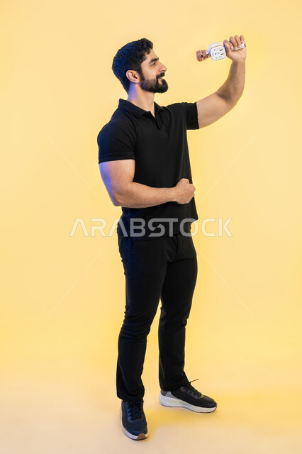 ممارسة هواية الغناء للتسلية والترفيه، الغناء بحماس وصوت عالي، الفن المسرحي الغنائي، تقديم العروضات المسرحية، بورتريه لشاب عربي خليجي اماراتي يرتدي الزي الكاجوال يحمل بيده الميكروفون، مواهب فنية في عالم الموسيقى، صورة الجسم بالطول الكامل، خلفية صفراء